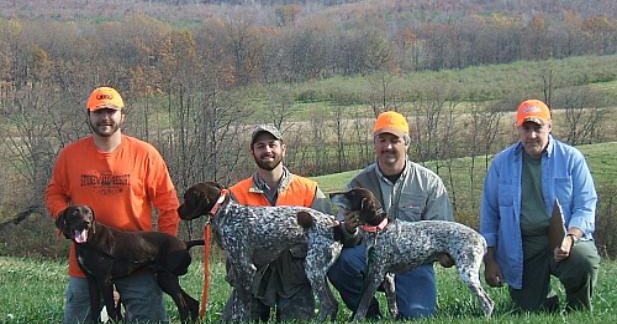 Four men wearing outdoor attire and bright orange caps pose with three dogs in a grassy area with a wooded backdrop. Two of the dogs are black-and-white, and the third is brown. The scene appears to be set in the countryside during autumn.