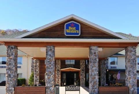 The image shows the front entrance of a Best Western hotel. The building features a wooden facade with stone pillars supporting a large overhang. The Best Western sign is prominently displayed above the entrance. An American flag is visible to the right of the entrance.