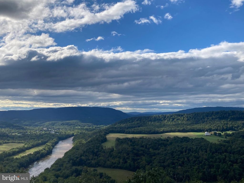 A scenic landscape showcases a valley with a winding river flanked by lush green hills under a partly cloudy sky. The distant horizon features rolling mountains, and patches of sunlight illuminate sections of the landscape. A few houses are scattered throughout the valley.