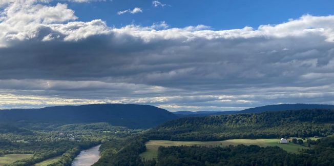 A scenic landscape showcases a valley with a winding river flanked by lush green hills under a partly cloudy sky. The distant horizon features rolling mountains, and patches of sunlight illuminate sections of the landscape. A few houses are scattered throughout the valley.