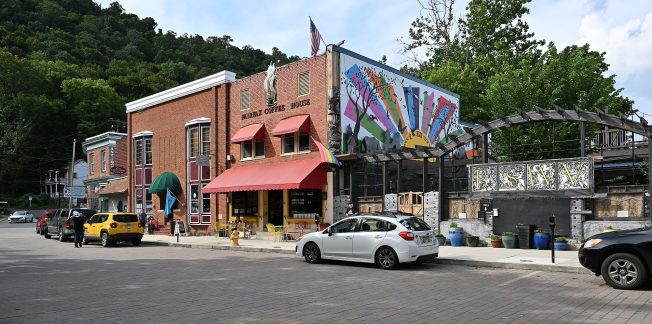 The image shows a vibrant street scene with several cars parked along a paved road. On the left, there's a red brick building with a green awning and a striped canopy, labeled 'Village Coffee House.' To the right, there's a building adorned with colorful street art depicting a hand holding paintbrushes. The setting has a quaint, small-town feel with lush green hills in the background.