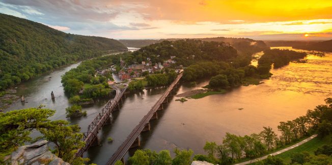 A scenic view of a river at sunset, with a bridge crossing over it and lush green hills on either side. A small town is nestled by the river, and the sky is glowing with warm orange and yellow hues. The landscape is bathed in the golden light of the setting sun.