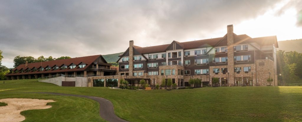 A wide-angle view of a large, multi-story stone and wood building with a red roof, situated on a well-maintained grassy area. The building has multiple windows, and the sun is partially obscured by clouds in the background. A path and sand area are visible in the foreground.