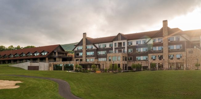 A wide-angle view of a large, multi-story stone and wood building with a red roof, situated on a well-maintained grassy area. The building has multiple windows, and the sun is partially obscured by clouds in the background. A path and sand area are visible in the foreground.