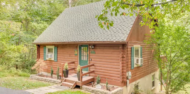 A small, charming wooden cabin with a pitched roof situated in a lush, wooded area. The cabin has a front porch with a staircase leading up to a green door adorned with a small decorative wreath. The surrounding greenery provides a serene, rustic atmosphere.