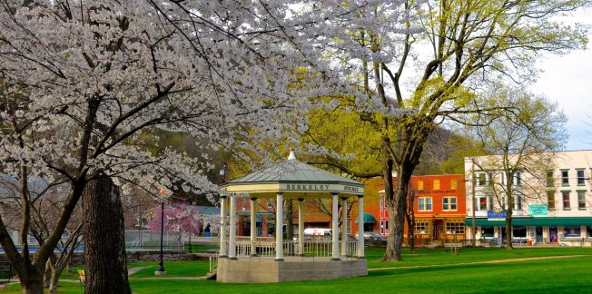 A charming park scene with a white gazebo under blossoming trees, surrounded by lush green grass. In the background, colorful buildings and tall trees with spring foliage are visible. The sky is partly cloudy, casting a serene ambiance over the park.