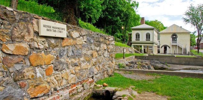 A stone wall with a plaque reading "George Washington's Bathtub (1769)" stands beside a small pool of water. In the background, a historical building with a green roof is surrounded by lush trees and grass under a partly cloudy sky.