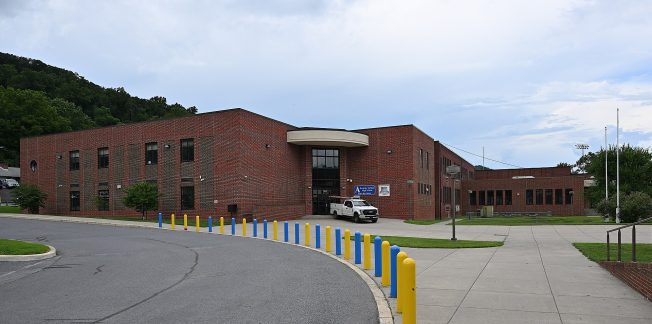 A modern, two-story red brick building with large windows and a central entrance, visible on a cloudy day. A white pickup truck is parked at the entrance. Brightly colored bollards line a curved driveway in the foreground with grassy areas on the sides.