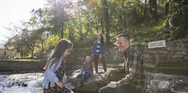 A family of four enjoys a sunny day at a park. A young girl plays near a small stream, while another girl sits on a rock. A woman stands in the background, and a man crouches near the stream, smiling. Trees and a sign reading "Thornhill Lord Forest Spring" are visible.