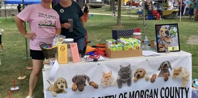 Two women standing behind a table under a white tent at an outdoor event. The table is covered with a banner that reads "HUMANE SOCIETY OF MORGAN COUNTY" and displays pet products, pamphlets, and a framed picture of a dog. Children and vendors are visible in the background.