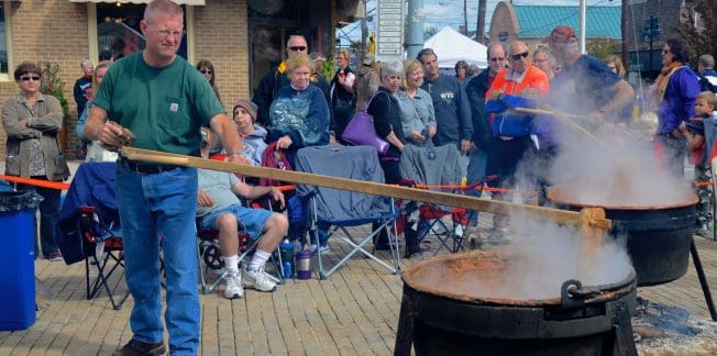 A man stirs contents in a large black kettle with a wooden stick at an outdoor event. Steam rises from the kettles, and a crowd of onlookers stands nearby. Some are seated in portable chairs, while others are standing, observing the cooking process.