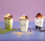 Three cups of layered sundaes are arranged in a row against a pastel purple background. Each sundae is topped with various toppings: the left one looks like it has matcha, the middle has caramel and nuts, and the right has fruit and chocolate crumbs.