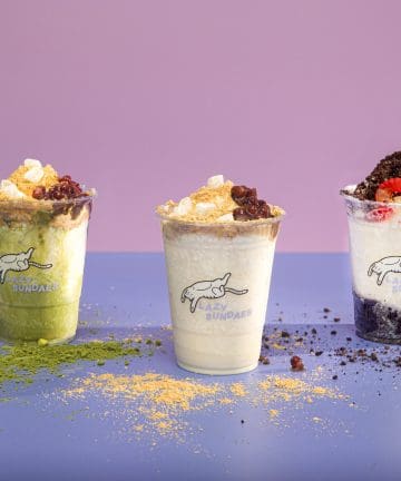 Three cups of layered sundaes are arranged in a row against a pastel purple background. Each sundae is topped with various toppings: the left one looks like it has matcha, the middle has caramel and nuts, and the right has fruit and chocolate crumbs.