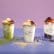 Three cups of layered sundaes are arranged in a row against a pastel purple background. Each sundae is topped with various toppings: the left one looks like it has matcha, the middle has caramel and nuts, and the right has fruit and chocolate crumbs.