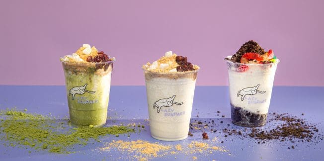 Three cups of layered sundaes are arranged in a row against a pastel purple background. Each sundae is topped with various toppings: the left one looks like it has matcha, the middle has caramel and nuts, and the right has fruit and chocolate crumbs.