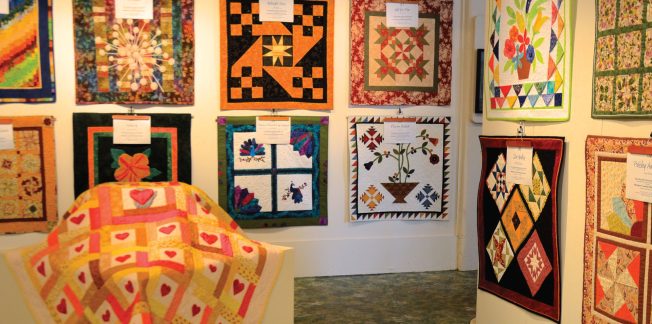 A display of colorful quilts hangs in a gallery, with intricate patterns and designs. Quilts feature floral, geometric, and heart-themed motifs. One quilt draped over a stand in the foreground showcases a heart design in pink and yellow tones. Tags describe each piece.