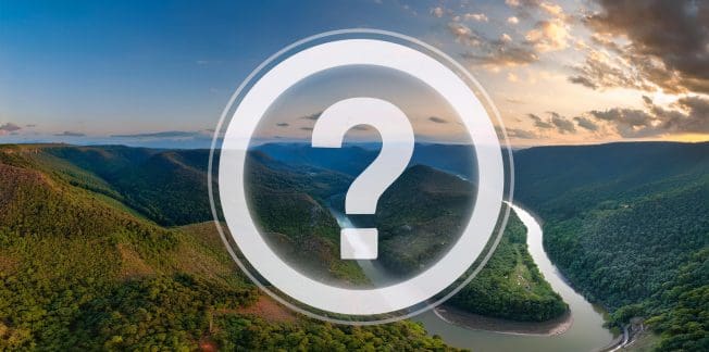 A panoramic view of a lush, green landscape featuring a winding river surrounded by forested hills. The sky above is partly cloudy with soft, golden light from the setting sun. A large, semi-transparent question mark icon is overlaid at the center of the image.