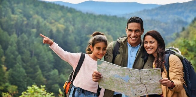 A family of three—two adults and a child—are outdoors in a forested mountainous area. The child is pointing into the distance while the adults hold a map, smiling. They are all wearing backpacks, suggesting they are on a hiking adventure.