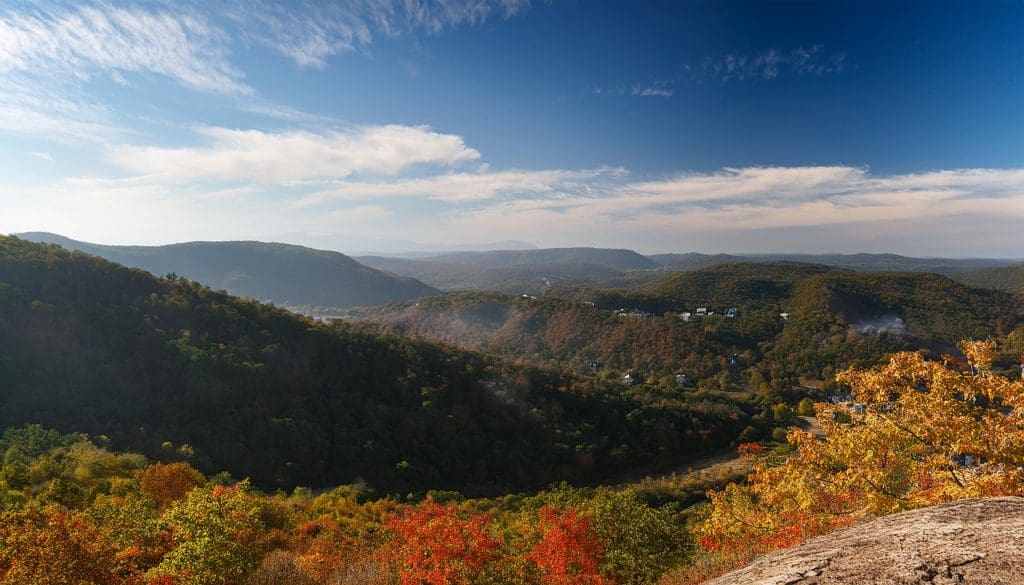 A panoramic view of a lush valley during autumn, displaying vibrant fall foliage in shades of red, orange, and yellow. Rolling hills extend into the distance under a clear blue sky with wispy white clouds. Small structures are scattered throughout the landscape.