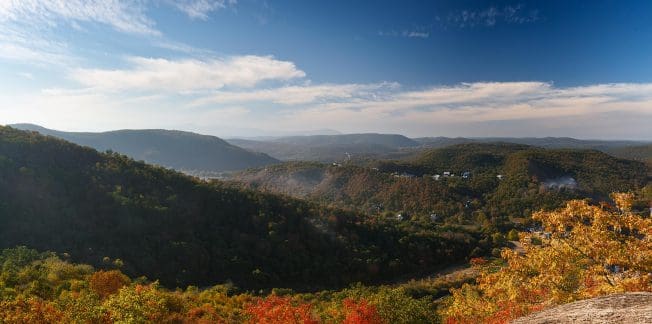 A panoramic view of a lush valley during autumn, displaying vibrant fall foliage in shades of red, orange, and yellow. Rolling hills extend into the distance under a clear blue sky with wispy white clouds. Small structures are scattered throughout the landscape.