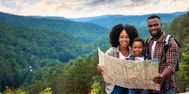 A family of three, consisting of a man, woman, and child, stands outdoors in a mountainous area holding a large map. The family appears happy and is dressed casually, suggesting they are on a hiking adventure. Lush green hills and a cloudy sky are visible in the background.