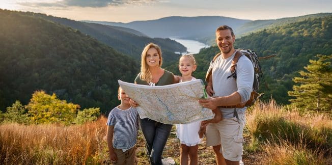 A smiling family of four stands on a hill with a scenic view of mountains, a river, and a forest. The parents hold a map while the children, one boy, and one girl, stand beside them. The father carries a backpack, indicating they are likely on a hiking trip.