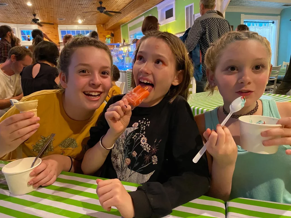 Three children sit at a table with white and green striped tablecloths, enjoying ice cream and popsicles. The child on the left is holding a cone, the child in the middle is holding a red popsicle, and the child on the right is eating ice cream with a spoon. They are smiling and appear to be having fun.