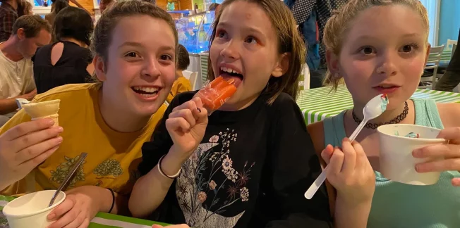 Three children sit at a table with white and green striped tablecloths, enjoying ice cream and popsicles. The child on the left is holding a cone, the child in the middle is holding a red popsicle, and the child on the right is eating ice cream with a spoon. They are smiling and appear to be having fun.