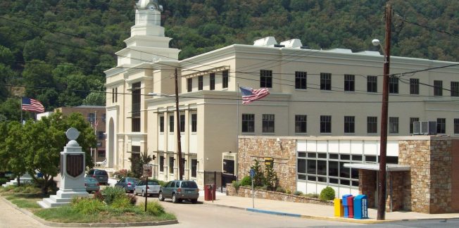 A beige, multi-story building with a clock tower stands near a wooded hill. American flags are seen on flagpoles. Several cars are parked nearby. The building is adjacent to a parking lot with a blue and yellow newspaper box. Power lines cross the scene.