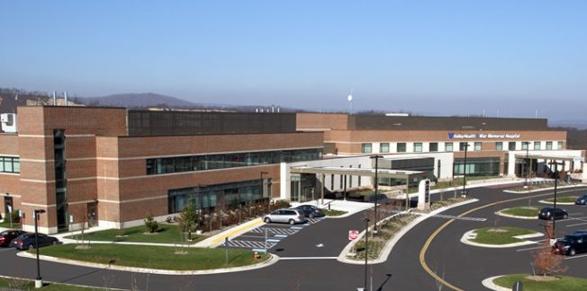 A modern brick and glass hospital building with an expansive parking area in the front, featuring multiple entry points and well-maintained landscaping. Several cars are parked nearby. The hospital is set against a backdrop of trees and distant hills.