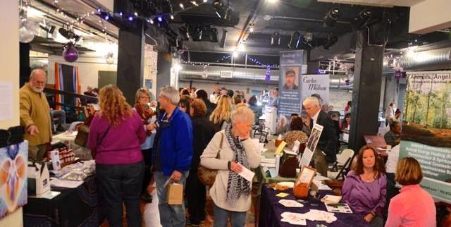 A busy indoor event with people browsing tables filled with brochures and products. The room has a festive atmosphere with string lights. Attendees are engaged in conversation and examining items on display.