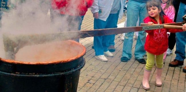 A young child in a red jacket helps stir a steaming pot outdoors. Several adults stand nearby, watching. The scene appears to be a community event on a brick-paved area.