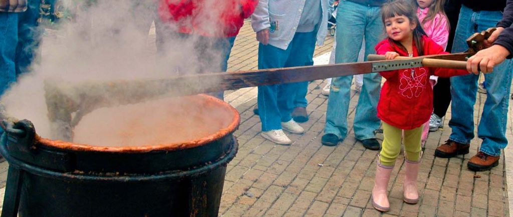 A young child in a red jacket helps stir a steaming pot outdoors. Several adults stand nearby, watching. The scene appears to be a community event on a brick-paved area.