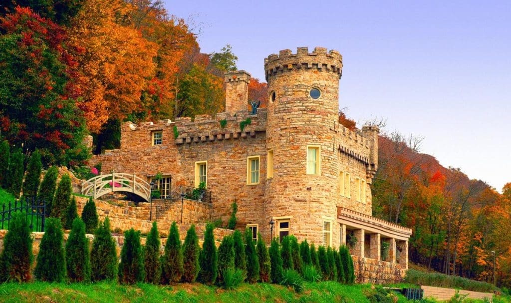 A picturesque stone castle surrounded by lush autumn foliage, featuring a rounded tower with crenellations, arched windows, and a small bridge in front. Trees display vibrant red, orange, and green hues under a clear blue sky.