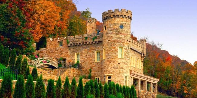 A picturesque stone castle surrounded by lush autumn foliage, featuring a rounded tower with crenellations, arched windows, and a small bridge in front. Trees display vibrant red, orange, and green hues under a clear blue sky.