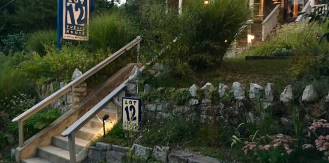 A cozy restaurant entrance with a lit sign reading "Lot 12 Public House." Stone steps are surrounded by lush green plants and a stone wall. The building has warm lights inside, creating an inviting atmosphere.