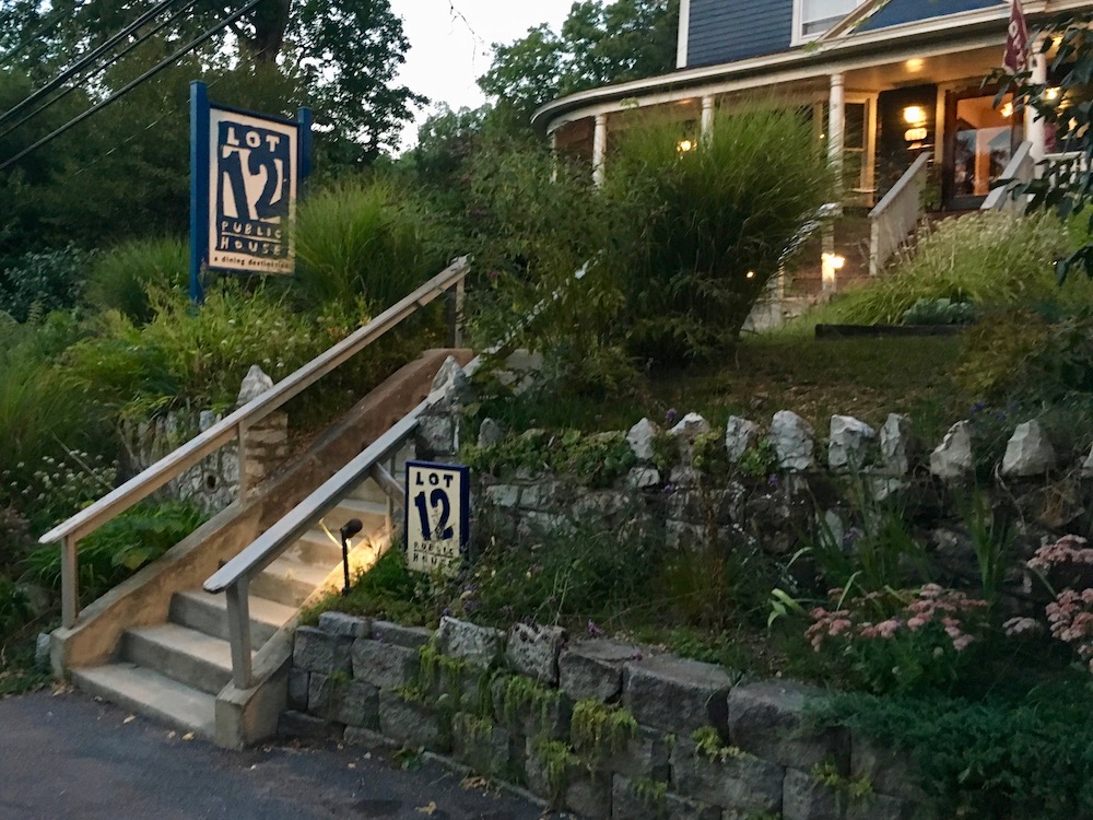 A cozy restaurant entrance with a lit sign reading "Lot 12 Public House." Stone steps are surrounded by lush green plants and a stone wall. The building has warm lights inside, creating an inviting atmosphere.