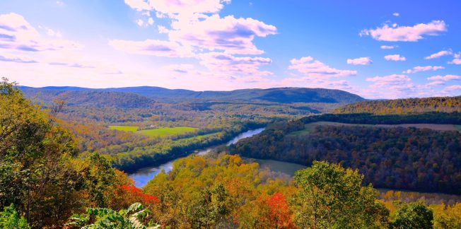 A scenic view of a winding river surrounded by vibrant autumn trees and rolling hills under a blue sky with scattered clouds. The landscape features lush greenery mixed with red, orange, and yellow foliage.